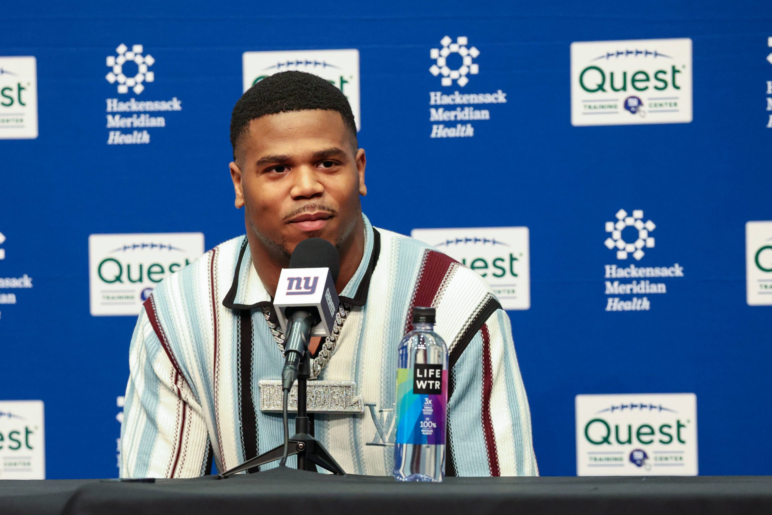 Abdul Carter sitzt an einem Tisch mit einem Mikrofon der New York Giants. Er trägt ein gestreiftes Hemd, das dezent auf seine Trikotnummer hindeutet, und eine große Halskette. Vor ihm steht eine Flasche LIFEWTR-Wasser. Im Hintergrund sind die Logos von Hackensack Meridian Health und Quest zu sehen. Diese Beschreibung wurde mit der FootballR KI automatisch generiert.