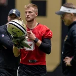 Tyler Shough in einem roten Trikot mit der Nummer 5, Gerüchten zufolge der neue Saints-Quarterback, hält drinnen einen Saints-Helm und ein Handtuch, während zwei Trainer – einer in schwarzer Saints-Ausrüstung, der andere mit Visier – aufmerksam zuschauen. Diese Beschreibung wurde mit der FootballR KI automatisch generiert.