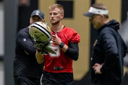 Tyler Shough in einem roten Trikot mit der Nummer 5, Gerüchten zufolge der neue Saints-Quarterback, hält drinnen einen Saints-Helm und ein Handtuch, während zwei Trainer – einer in schwarzer Saints-Ausrüstung, der andere mit Visier – aufmerksam zuschauen. Diese Beschreibung wurde mit der FootballR KI automatisch generiert.