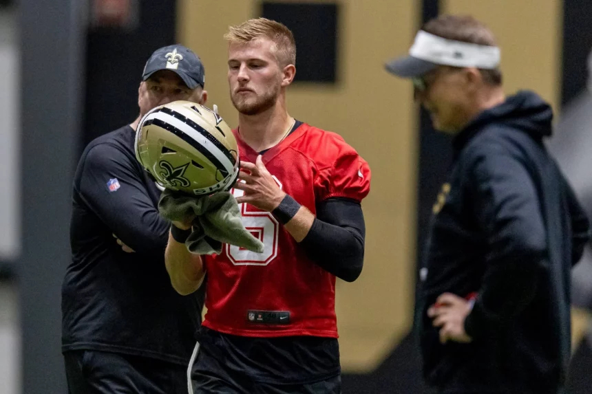 Tyler Shough in einem roten Trikot mit der Nummer 5, Gerüchten zufolge der neue Saints-Quarterback, hält drinnen einen Saints-Helm und ein Handtuch, während zwei Trainer – einer in schwarzer Saints-Ausrüstung, der andere mit Visier – aufmerksam zuschauen. Diese Beschreibung wurde mit der FootballR KI automatisch generiert.