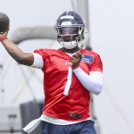 Cam Ward in rotem Trainingstrikot und blauem Helm, der Führungsqualitäten zeigt, bereitet sich beim Outdoor-Training darauf vor, mit der rechten Hand einen Football zu werfen. Er trägt einen weißen Armstulpen und ist mitten in der Bewegung, fokussiert auf sein Ziel. Im Hintergrund sind Ausrüstung und Himmel unscharf. Diese Beschreibung wurde mit der FootballR KI automatisch generiert.