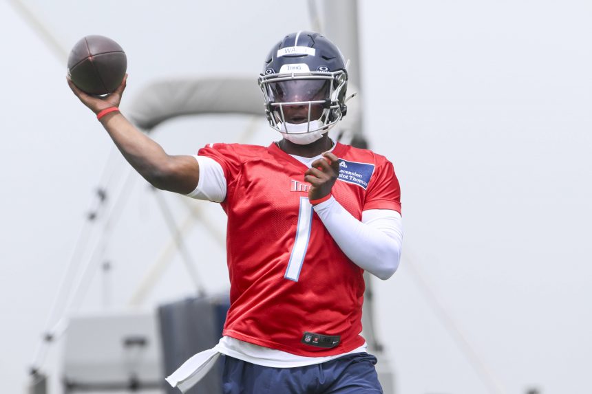 Cam Ward in rotem Trainingstrikot und blauem Helm, der Führungsqualitäten zeigt, bereitet sich beim Outdoor-Training darauf vor, mit der rechten Hand einen Football zu werfen. Er trägt einen weißen Armstulpen und ist mitten in der Bewegung, fokussiert auf sein Ziel. Im Hintergrund sind Ausrüstung und Himmel unscharf. Diese Beschreibung wurde mit der FootballR KI automatisch generiert.