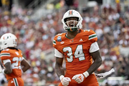 Ein Footballspieler der Miami Hurricanes, Adarius Hayes, steht mit geballten Fäusten auf dem Spielfeld und jubelt laut. Im Hintergrund sind ein weiterer Spieler und eine verschwommene Menge zu sehen – Adarius Hayes Unfall macht Schlagzeilen. Diese Beschreibung wurde mit der FootballR KI automatisch generiert.