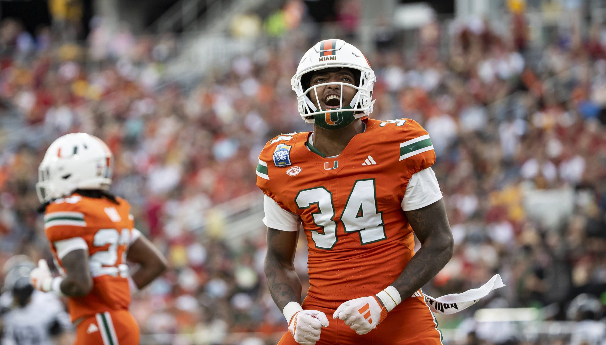 Ein Footballspieler der Miami Hurricanes, Adarius Hayes, steht mit geballten Fäusten auf dem Spielfeld und jubelt laut. Im Hintergrund sind ein weiterer Spieler und eine verschwommene Menge zu sehen – Adarius Hayes Unfall macht Schlagzeilen. Diese Beschreibung wurde mit der FootballR KI automatisch generiert.