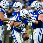 Footballspieler der Indianapolis Colts in blau-weißen Trikots feiern auf dem Spielfeld. Ein Spieler der Indianapolis Colts hält den Ball und ist während des Spiels von Teamkollegen umringt, die ihm gratulieren. Im unscharfen Hintergrund ist eine jubelnde Menge zu sehen. Diese Beschreibung wurde mit der FootballR KI automatisch generiert.