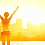 Eine Frau steht bei Sonnenaufgang mit triumphierend erhobenen Armen im Freien und umarmt ein gesünderes Leben. Sie blickt auf eine helle Stadtsilhouette, das Sonnenlicht lässt sie golden leuchten. Sie ist sportlich gekleidet und strahlt sommerliche Energie aus - ein echter Sommer-Guide für ein gesundes Leben. Diese Beschreibung wurde mit der FootballR KI automatisch generiert.