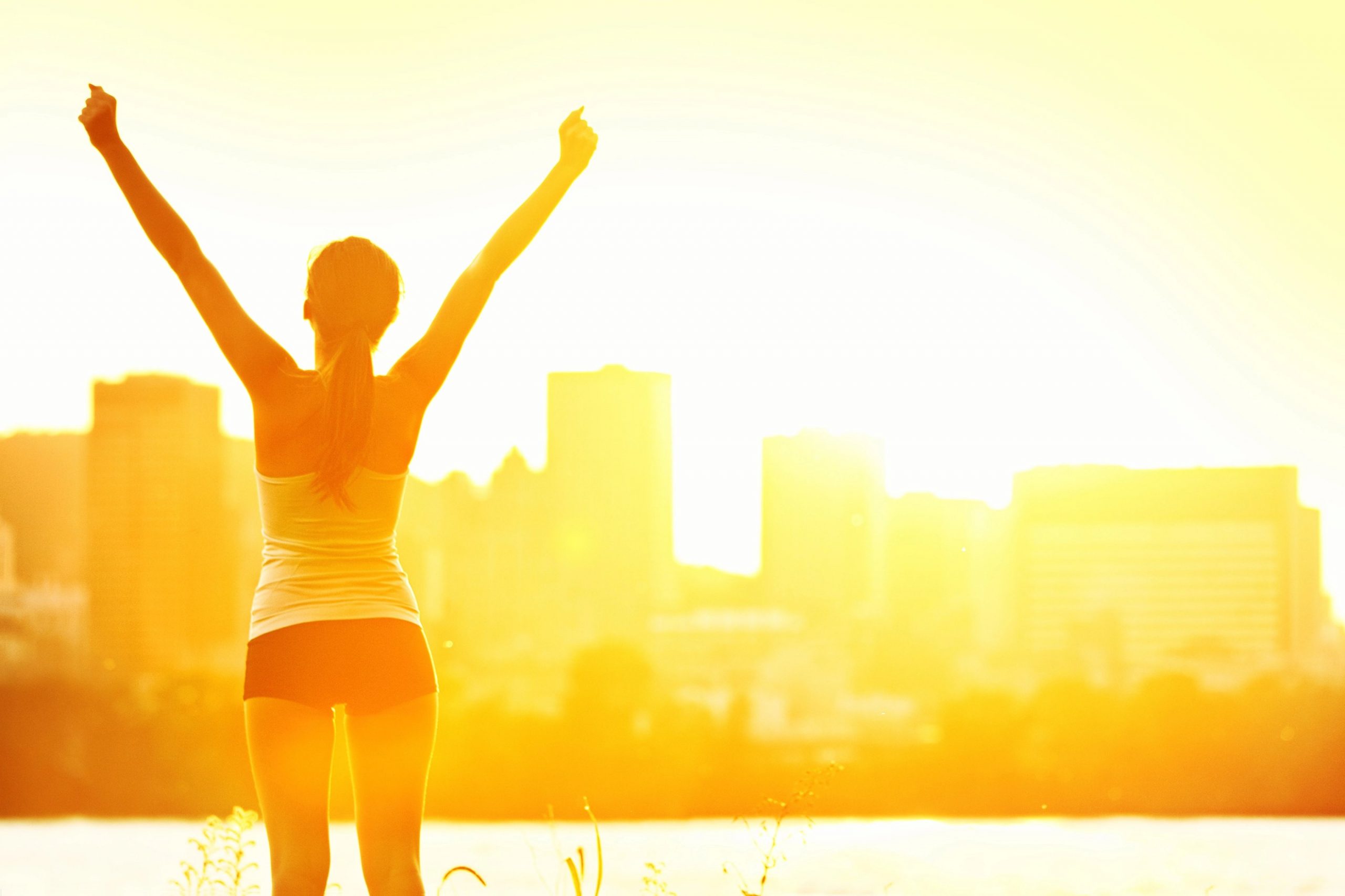 Eine Frau steht bei Sonnenaufgang mit triumphierend erhobenen Armen im Freien und umarmt ein gesünderes Leben. Sie blickt auf eine helle Stadtsilhouette, das Sonnenlicht lässt sie golden leuchten. Sie ist sportlich gekleidet und strahlt sommerliche Energie aus - ein echter Sommer-Guide für ein gesundes Leben. Diese Beschreibung wurde mit der FootballR KI automatisch generiert.