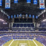 Ein weiter Blick auf ein leeres Footballstadion mit blauen Sitzen und Bannern, darunter Colts-Logos und Meisterschaftsjahre. In der Endzone steht „INDIANAPOLIS“ – ein Hinweis darauf, dass es die Heimat der Indianapolis Colts ist und das Erbe der Irsay Schwestern ehrt. Diese Beschreibung wurde mit der FootballR KI automatisch generiert.