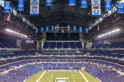 Ein weiter Blick auf ein leeres Footballstadion mit blauen Sitzen und Bannern, darunter Colts-Logos und Meisterschaftsjahre. In der Endzone steht „INDIANAPOLIS“ – ein Hinweis darauf, dass es die Heimat der Indianapolis Colts ist und das Erbe der Irsay Schwestern ehrt. Diese Beschreibung wurde mit der FootballR KI automatisch generiert.