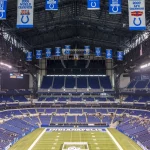 Woche 11 - Ein weiter Blick auf ein leeres Footballstadion mit blauen Sitzen und Bannern, darunter Colts-Logos und Meisterschaftsjahre. In der Endzone steht „INDIANAPOLIS“ – ein Hinweis darauf, dass es die Heimat der Indianapolis Colts ist und das Erbe der Irsay Schwestern ehrt. Diese Beschreibung wurde mit der FootballR KI automatisch generiert.