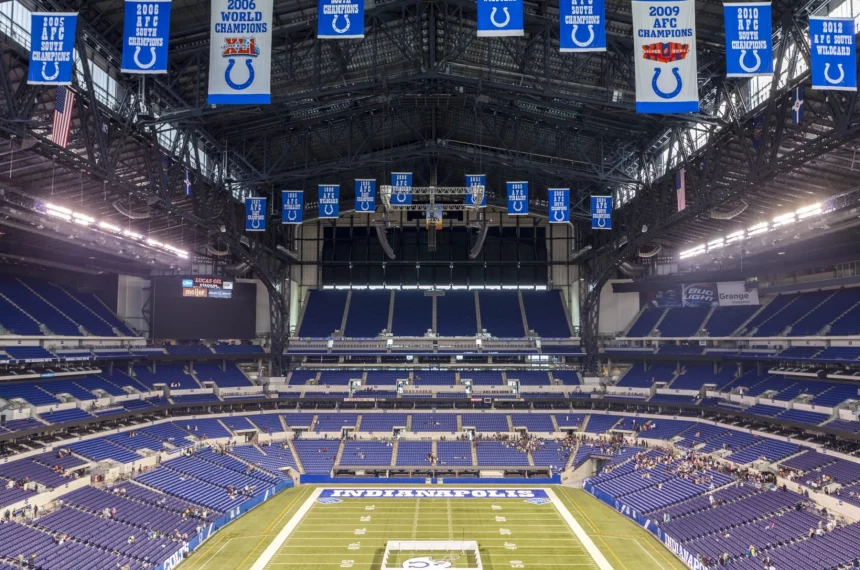 Woche 11 - Ein weiter Blick auf ein leeres Footballstadion mit blauen Sitzen und Bannern, darunter Colts-Logos und Meisterschaftsjahre. In der Endzone steht „INDIANAPOLIS“ – ein Hinweis darauf, dass es die Heimat der Indianapolis Colts ist und das Erbe der Irsay Schwestern ehrt. Diese Beschreibung wurde mit der FootballR KI automatisch generiert.
