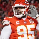Chiefs und Royals - Ein Footballspieler im roten Trikot der Kansas City Chiefs (Nummer 95) hebt seine linke Hand ans Ohr, als würde er zuhören. Die verschwommene Stadionmenge deutet auf große Stadionprojekte hin. In der Ecke ist die Umrisslinie des Staates Missouri mit einem Stern und dem Schriftzug „MISSOURI“ zu sehen. Diese Beschreibung wurde mit der FootballR KI automatisch generiert.