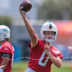 Ein Footballspieler in einem roten Trikot und Helm der Miami Dolphins, der Zach Wilson ähnelt, wirft beim Training einen Football. Ein weiterer Spieler steht hinter ihm. Eine gepunktete Linie zeigt am unteren Bildrand eine Flugroute von New York nach Florida. Diese Beschreibung wurde mit der FootballR KI automatisch generiert.