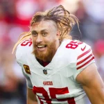 Ein Footballspieler, George Kittle, mit langen blonden Haaren und Bart, der ein weißes Trikot mit roten Streifen und der Nummer 85 trägt, schreit aufgeregt auf dem Feld – vielleicht feiert er Tight-End-Gehälter. In der Ecke ist der Umriss von Kalifornien mit einem Stern zu sehen. Diese Beschreibung wurde mit der FootballR KI automatisch generiert.