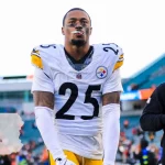 DeShon Elliott, Footballspieler der Pittsburgh Steelers, mit der Trikotnummer 25, steht auf dem Spielfeld und blickt nach oben. Ein weiterer maskierter Steelers-Spieler ist in der Nähe. Im Hintergrund sind Stadionsitze mit einer Overlay-Grafik von Pennsylvania und einem Stecknadelsymbol zu sehen. Diese Beschreibung wurde mit der FootballR KI automatisch generiert.