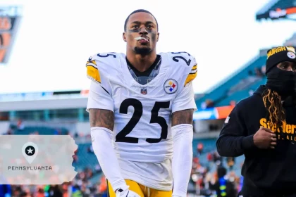 DeShon Elliott, Footballspieler der Pittsburgh Steelers, mit der Trikotnummer 25, steht auf dem Spielfeld und blickt nach oben. Ein weiterer maskierter Steelers-Spieler ist in der Nähe. Im Hintergrund sind Stadionsitze mit einer Overlay-Grafik von Pennsylvania und einem Stecknadelsymbol zu sehen. Diese Beschreibung wurde mit der FootballR KI automatisch generiert.
