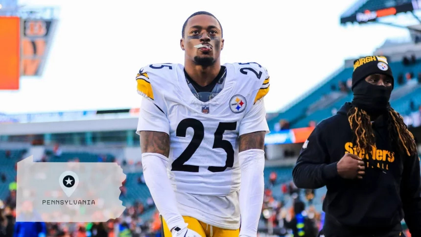 DeShon Elliott, Footballspieler der Pittsburgh Steelers, mit der Trikotnummer 25, steht auf dem Spielfeld und blickt nach oben. Ein weiterer maskierter Steelers-Spieler ist in der Nähe. Im Hintergrund sind Stadionsitze mit einer Overlay-Grafik von Pennsylvania und einem Stecknadelsymbol zu sehen. Diese Beschreibung wurde mit der FootballR KI automatisch generiert.