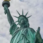 Football x Football - Eine Nahaufnahme der Freiheitsstatue vor einem bewölkten Himmel. Ihre erhobene Fackel und Krone sind sichtbar. Der handgeschriebene Text „New York City x Football“ erscheint in Schwarz auf der linken Seite des Bildes. Diese Beschreibung wurde mit der FootballR KI automatisch generiert.