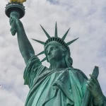 Football x Football - Eine Nahaufnahme der Freiheitsstatue vor einem bewölkten Himmel. Ihre erhobene Fackel und Krone sind sichtbar. Der handgeschriebene Text „New York City x Football“ erscheint in Schwarz auf der linken Seite des Bildes. Diese Beschreibung wurde mit der FootballR KI automatisch generiert.