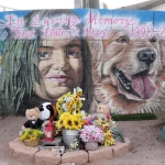 Ein Wandgemälde an einer Backsteinmauer zeigt ein Mädchen und einen Golden Retriever. Darüber steht „In liebevoller Erinnerung an Tina Tintor und Max 1998–2021“ – eine herzliche Hommage an die Tragödie um Henry Ruggs. Darunter befinden sich Blumen, Stofftiere, Kerzen und ein Foto. Unten links ist ein Nevada-Grabstein zu sehen. Diese Beschreibung wurde mit der FootballR KI automatisch generiert.