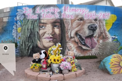 Ein Wandgemälde an einer Backsteinmauer zeigt ein Mädchen und einen Golden Retriever. Darüber steht „In liebevoller Erinnerung an Tina Tintor und Max 1998–2021“ – eine herzliche Hommage an die Tragödie um Henry Ruggs. Darunter befinden sich Blumen, Stofftiere, Kerzen und ein Foto. Unten links ist ein Nevada-Grabstein zu sehen. Diese Beschreibung wurde mit der FootballR KI automatisch generiert.