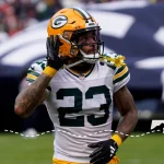 Ein Footballspieler der Green Bay Packers, Jaire Alexander, im Trikot mit der Nummer 23, hält sich auf dem Spielfeld das Ohr zu. Darunter verbindet eine gestrichelte Linie ein Symbol von Wisconsin mit einem Symbol von Maryland, was auf eine Reise zwischen den beiden Staaten hinweist. Diese Beschreibung wurde mit der FootballR KI automatisch generiert.