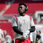 Ein Footballspieler, Antonio Brown, trägt ein graues Trikot, rote Shorts, weiße Handschuhe und große Kopfhörer trägt, steht auf einem fast leeren Feld. Konzentriert blickt er zur Seite, während sich im Hintergrund andere Spieler aufwärmen. Diese Beschreibung wurde mit der FootballR KI automatisch generiert.