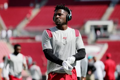 Ein Footballspieler, Antonio Brown, trägt ein graues Trikot, rote Shorts, weiße Handschuhe und große Kopfhörer trägt, steht auf einem fast leeren Feld. Konzentriert blickt er zur Seite, während sich im Hintergrund andere Spieler aufwärmen. Diese Beschreibung wurde mit der FootballR KI automatisch generiert.