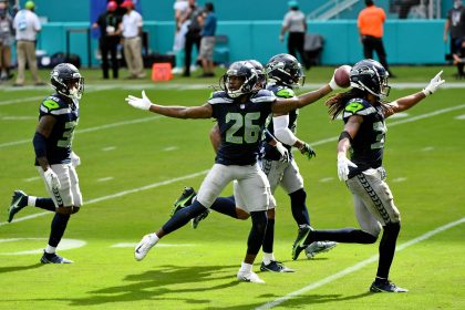 Vier Footballspieler der Seattle Seahawks, darunter Shaquill Griffin in dunkelblauen Trikots, feiern auf einem sonnigen Feld. Ein Spieler im Vordergrund hält den Football in die Höhe, während ein anderer seine Arme weit ausbreitet, während sie gemeinsam laufen. Andere Spieler und Zuschauer sind im Hintergrund zu sehen. Diese Beschreibung wurde mit der FootballR KI automatisch generiert.