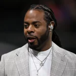 Ein Mann mit langen Zöpfen und Bart, der Richard Sherman von den Seahawks ähnelt, trägt ein hellgraues kariertes Jackett über einem weißen Hemd und mehrere Halsketten. Drinnen blickt er zur Seite, neben ihm ist eine andere Person teilweise sichtbar. Diese Beschreibung wurde mit der FootballR KI automatisch generiert.