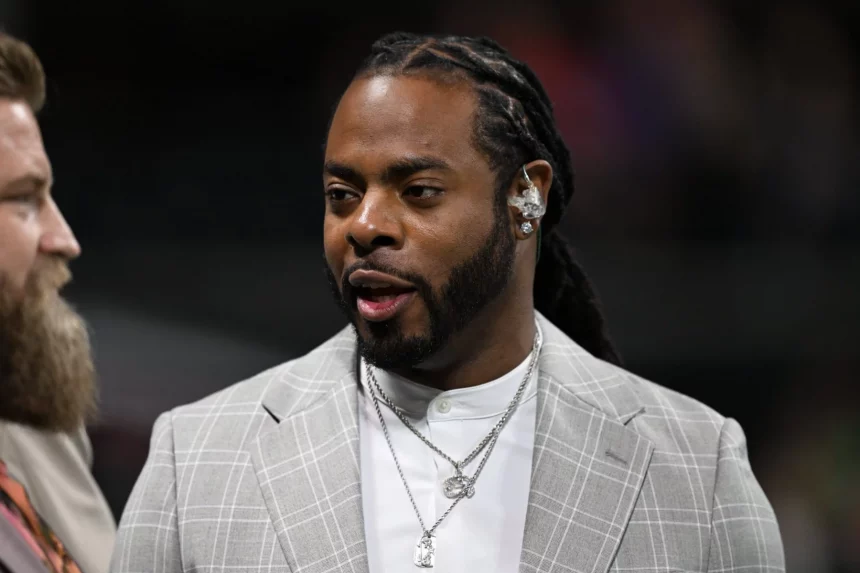 Ein Mann mit langen Zöpfen und Bart, der Richard Sherman von den Seahawks ähnelt, trägt ein hellgraues kariertes Jackett über einem weißen Hemd und mehrere Halsketten. Drinnen blickt er zur Seite, neben ihm ist eine andere Person teilweise sichtbar. Diese Beschreibung wurde mit der FootballR KI automatisch generiert.