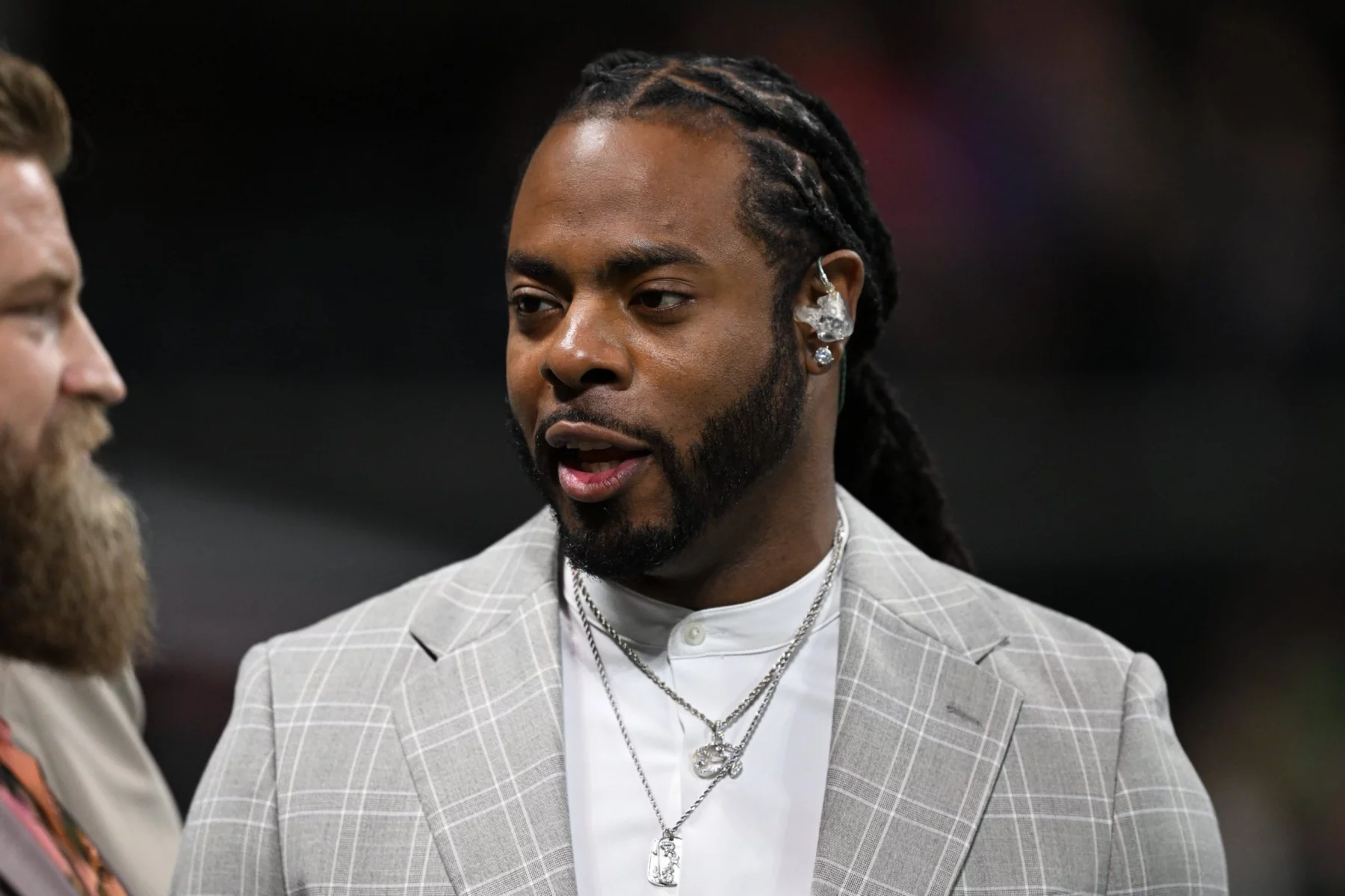 Ein Mann mit langen Zöpfen und Bart, der Richard Sherman von den Seahawks ähnelt, trägt ein hellgraues kariertes Jackett über einem weißen Hemd und mehrere Halsketten. Drinnen blickt er zur Seite, neben ihm ist eine andere Person teilweise sichtbar. Diese Beschreibung wurde mit der FootballR KI automatisch generiert.