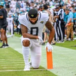 Gabe Davis, in einer weißen Uniform der Jacksonville Jaguars, kniet in der Nähe des orangefarbenen Pylonen am Rand des Spielfelds, den Kopf gesenkt und die Hand auf dem Boden, während im Hintergrund unscharf eine Menge, Schiedsrichter und Mannschaftsmitarbeiter zu sehen sind. Diese Beschreibung wurde mit der FootballR KI automatisch generiert.