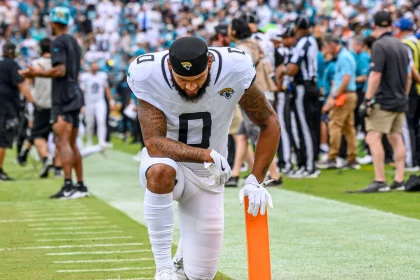Gabe Davis, in einer weißen Uniform der Jacksonville Jaguars, kniet in der Nähe des orangefarbenen Pylonen am Rand des Spielfelds, den Kopf gesenkt und die Hand auf dem Boden, während im Hintergrund unscharf eine Menge, Schiedsrichter und Mannschaftsmitarbeiter zu sehen sind. Diese Beschreibung wurde mit der FootballR KI automatisch generiert.