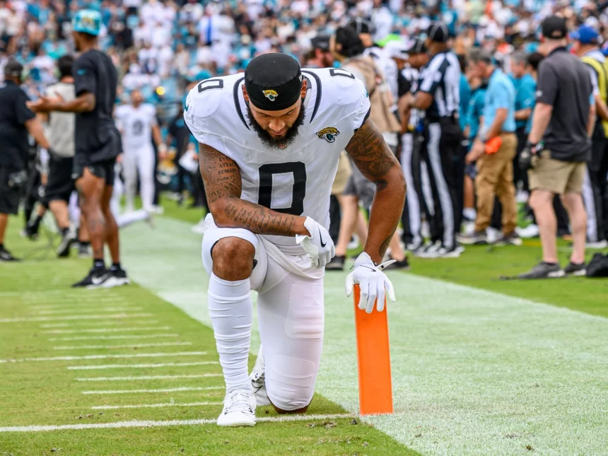 Gabe Davis, in einer weißen Uniform der Jacksonville Jaguars, kniet in der Nähe des orangefarbenen Pylonen am Rand des Spielfelds, den Kopf gesenkt und die Hand auf dem Boden, während im Hintergrund unscharf eine Menge, Schiedsrichter und Mannschaftsmitarbeiter zu sehen sind. Diese Beschreibung wurde mit der FootballR KI automatisch generiert.