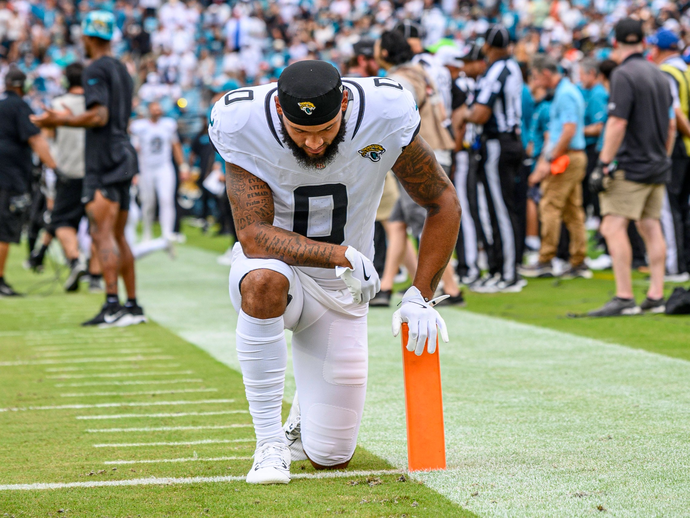 Gabe Davis, in einer weißen Uniform der Jacksonville Jaguars, kniet in der Nähe des orangefarbenen Pylonen am Rand des Spielfelds, den Kopf gesenkt und die Hand auf dem Boden, während im Hintergrund unscharf eine Menge, Schiedsrichter und Mannschaftsmitarbeiter zu sehen sind. Diese Beschreibung wurde mit der FootballR KI automatisch generiert.