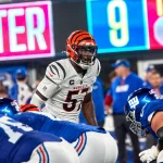 Germaine Pratt, ein Footballspieler der Cincinnati Bengals in weißer Uniform und orange gestreiftem Helm, steht der Offensive Line der New York Giants in blauen Uniformen gegenüber und bereitet sich auf einen Spielzug vor. Im Hintergrund sind Trainer, eine digitale Anzeigetafel und Stadionlichter zu sehen. Diese Beschreibung wurde mit der FootballR KI automatisch generiert.