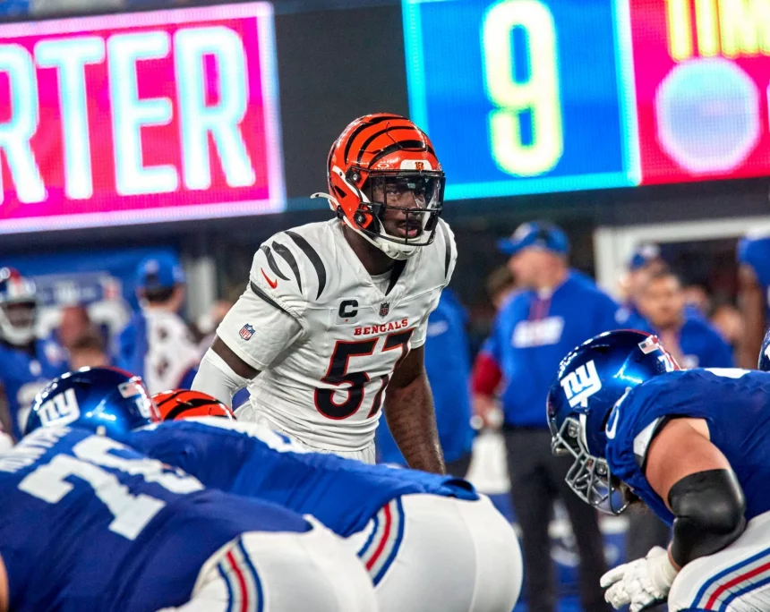 Germaine Pratt, ein Footballspieler der Cincinnati Bengals in weißer Uniform und orange gestreiftem Helm, steht der Offensive Line der New York Giants in blauen Uniformen gegenüber und bereitet sich auf einen Spielzug vor. Im Hintergrund sind Trainer, eine digitale Anzeigetafel und Stadionlichter zu sehen. Diese Beschreibung wurde mit der FootballR KI automatisch generiert.