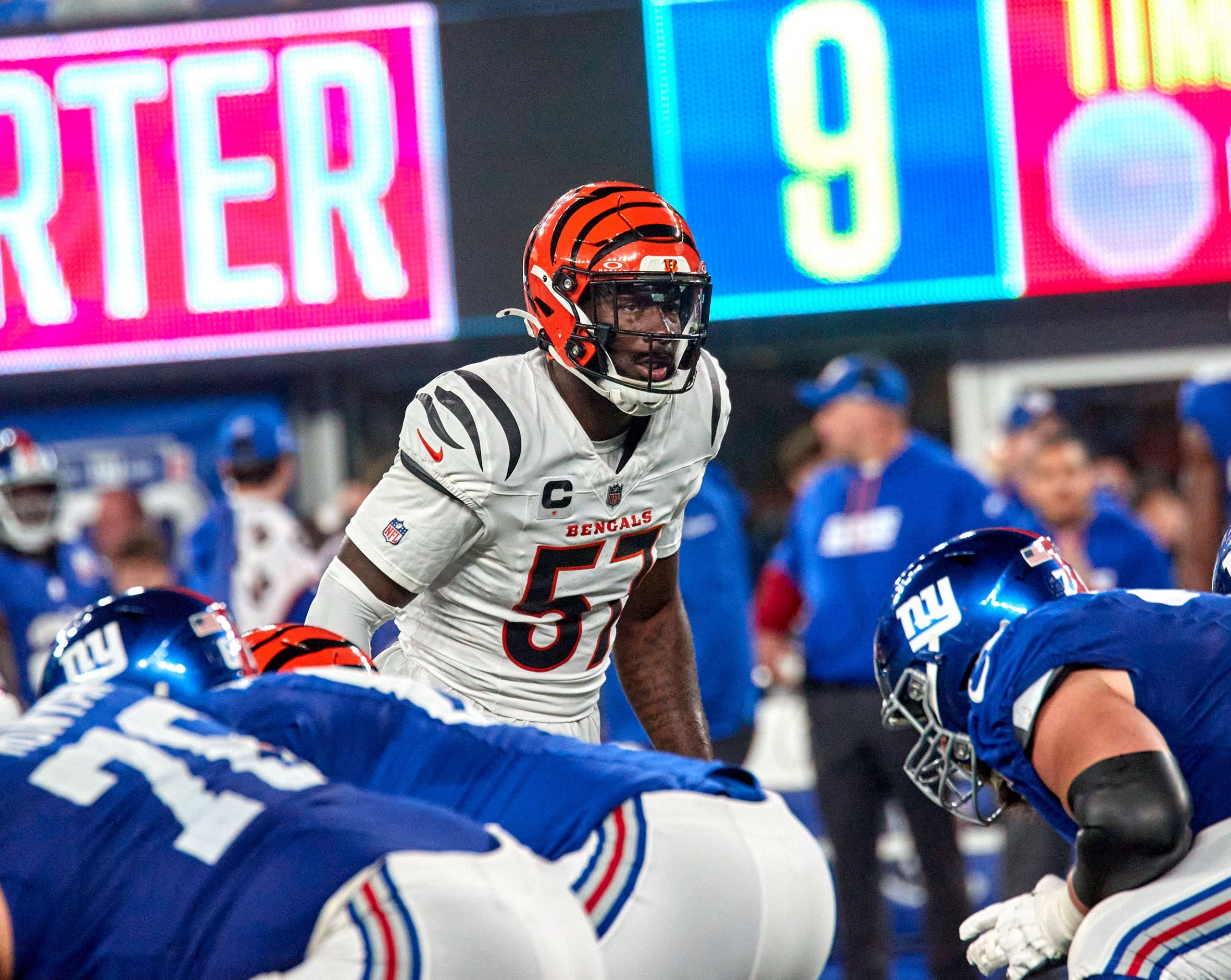 Germaine Pratt, ein Footballspieler der Cincinnati Bengals in weißer Uniform und orange gestreiftem Helm, steht der Offensive Line der New York Giants in blauen Uniformen gegenüber und bereitet sich auf einen Spielzug vor. Im Hintergrund sind Trainer, eine digitale Anzeigetafel und Stadionlichter zu sehen. Diese Beschreibung wurde mit der FootballR KI automatisch generiert.