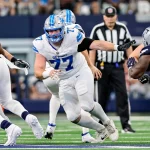 Frank Ragnow, Nummer 77 der Detroit Lions in weißer Uniform mit blauen Akzenten, blockt einen Spieler der Dallas Cowboys, Nummer 58, in marineblauer Uniform. Im Hintergrund sind weitere Spieler und ein Schiedsrichter auf dem Footballfeld zu sehen. Diese Beschreibung wurde mit der FootballR KI automatisch generiert.