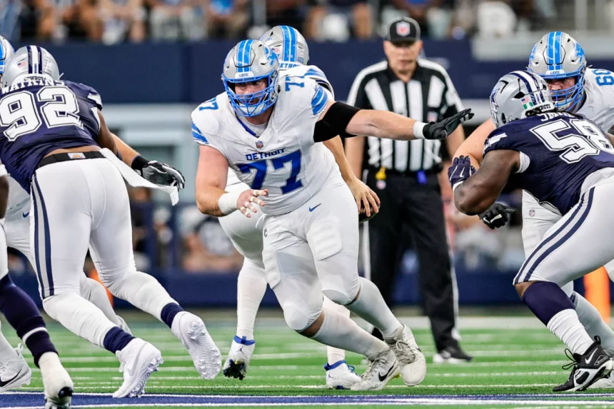 Frank Ragnow, Nummer 77 der Detroit Lions in weißer Uniform mit blauen Akzenten, blockt einen Spieler der Dallas Cowboys, Nummer 58, in marineblauer Uniform. Im Hintergrund sind weitere Spieler und ein Schiedsrichter auf dem Footballfeld zu sehen. Diese Beschreibung wurde mit der FootballR KI automatisch generiert.