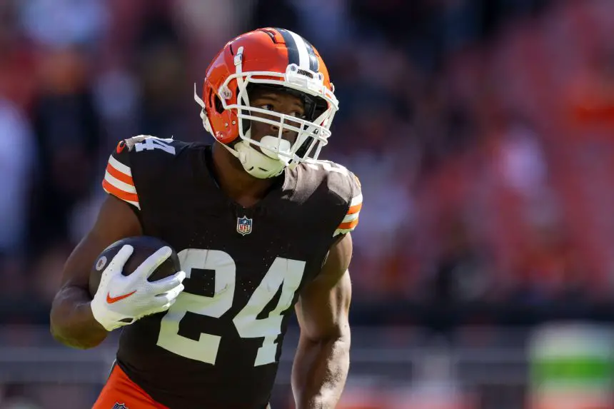 Nick Chubb trägt ein braunes Cleveland Browns-Trikot mit der Nummer 24 und läuft mit einem Football in der Hand. Er trägt einen Helm und weiße Handschuhe, während das Sonnenlicht ihn vor dem verschwommenen Stadionhintergrund hervorhebt. Diese Beschreibung wurde mit der FootballR KI automatisch generiert.
