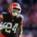 Nick Chubb trägt ein braunes Cleveland Browns-Trikot mit der Nummer 24 und läuft mit einem Football in der Hand. Er trägt einen Helm und weiße Handschuhe, während das Sonnenlicht ihn vor dem verschwommenen Stadionhintergrund hervorhebt. Diese Beschreibung wurde mit der FootballR KI automatisch generiert.
