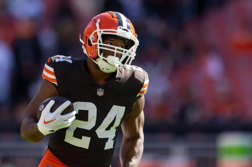 Nick Chubb trägt ein braunes Cleveland Browns-Trikot mit der Nummer 24 und läuft mit einem Football in der Hand. Er trägt einen Helm und weiße Handschuhe, während das Sonnenlicht ihn vor dem verschwommenen Stadionhintergrund hervorhebt. Diese Beschreibung wurde mit der FootballR KI automatisch generiert.