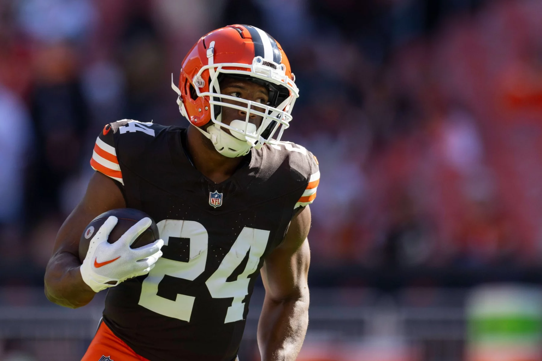 Nick Chubb trägt ein braunes Cleveland Browns-Trikot mit der Nummer 24 und läuft mit einem Football in der Hand. Er trägt einen Helm und weiße Handschuhe, während das Sonnenlicht ihn vor dem verschwommenen Stadionhintergrund hervorhebt. Diese Beschreibung wurde mit der FootballR KI automatisch generiert.