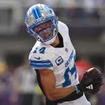 Amon-Ra St. Brown, ein Footballspieler der Detroit Lions in weiß-blauer Uniform mit der Nummer 14, hält während eines Spiels einen Football und blickt über die Schulter zurück. Er trägt einen silbernen Helm mit blauem Gesichtsschutz und ist mitten im Schritt auf dem Spielfeld. Diese Beschreibung wurde mit der FootballR KI automatisch generiert.