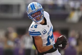 Amon-Ra St. Brown, ein Footballspieler der Detroit Lions in weiß-blauer Uniform mit der Nummer 14, hält während eines Spiels einen Football und blickt über die Schulter zurück. Er trägt einen silbernen Helm mit blauem Gesichtsschutz und ist mitten im Schritt auf dem Spielfeld. Diese Beschreibung wurde mit der FootballR KI automatisch generiert.