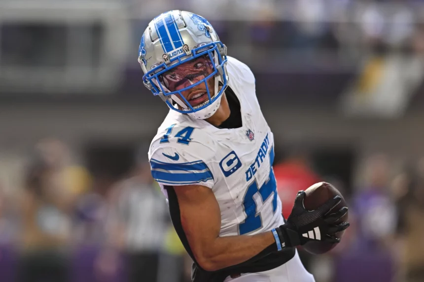 Amon-Ra St. Brown, ein Footballspieler der Detroit Lions in weiß-blauer Uniform mit der Nummer 14, hält während eines Spiels einen Football und blickt über die Schulter zurück. Er trägt einen silbernen Helm mit blauem Gesichtsschutz und ist mitten im Schritt auf dem Spielfeld. Diese Beschreibung wurde mit der FootballR KI automatisch generiert.