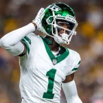 Sauce Gardner und Garrett Wilson - Ein Footballspieler in weiß-grüner New York Jets-Uniform und Helm, der mit der rechten Hand am Ohr jubelt, reagiert auf spannende Vertragsverlängerungen. Er trägt die Nummer 1 und steht auf dem Spielfeld, im Hintergrund ist eine unscharfe Menschenmenge zu sehen. Diese Beschreibung wurde mit der FootballR KI automatisch generiert.