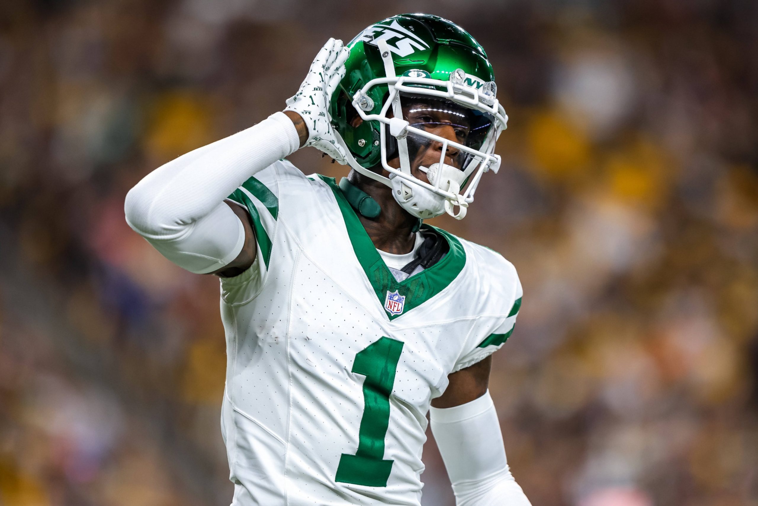 Sauce Gardner und Garrett Wilson - Ein Footballspieler in weiß-grüner New York Jets-Uniform und Helm, der mit der rechten Hand am Ohr jubelt, reagiert auf spannende Vertragsverlängerungen. Er trägt die Nummer 1 und steht auf dem Spielfeld, im Hintergrund ist eine unscharfe Menschenmenge zu sehen. Diese Beschreibung wurde mit der FootballR KI automatisch generiert.